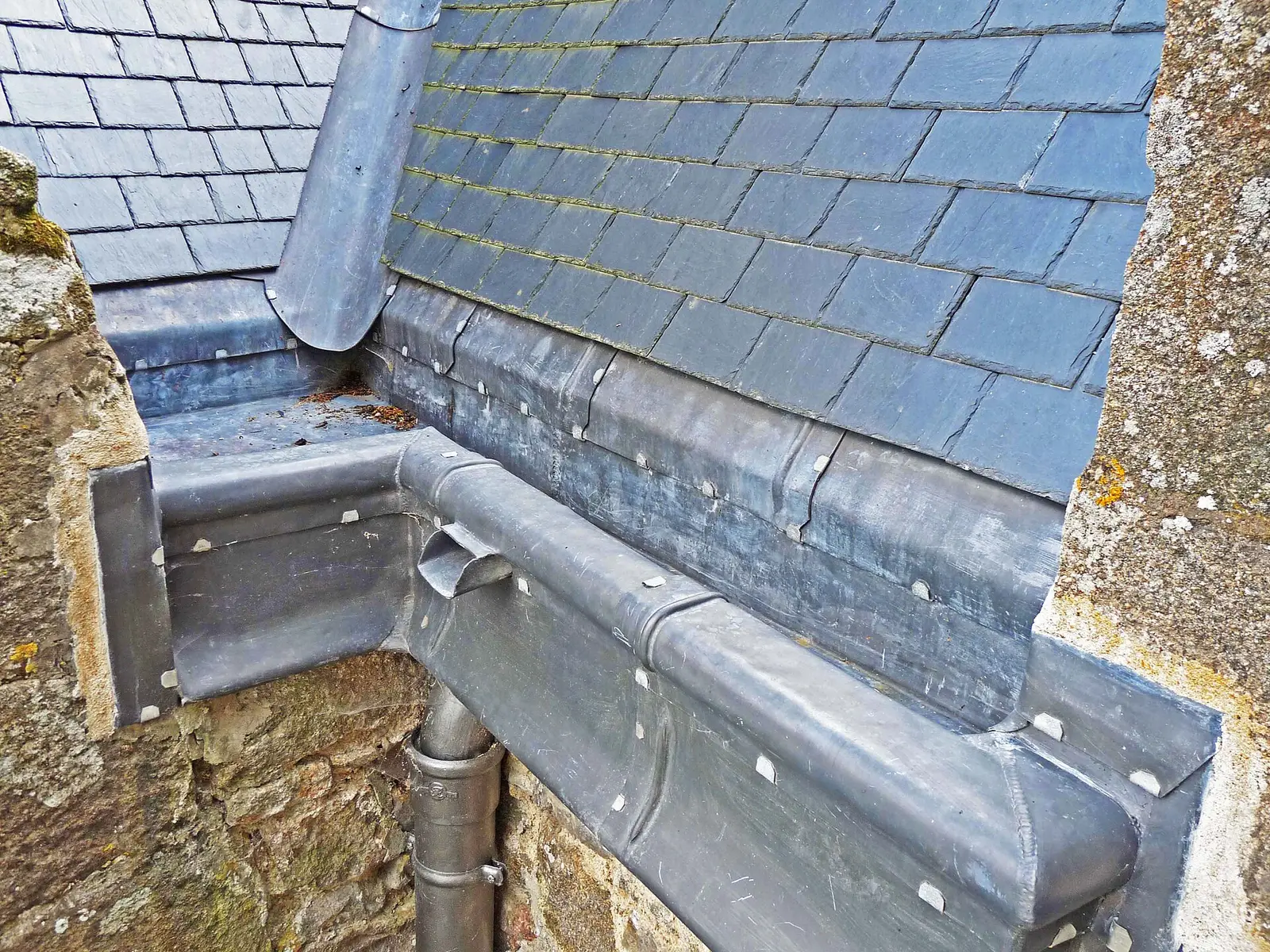 Metal roof gutter running along the bottom of a slate roof, with a downspout outlet dropping into a round drainpipe