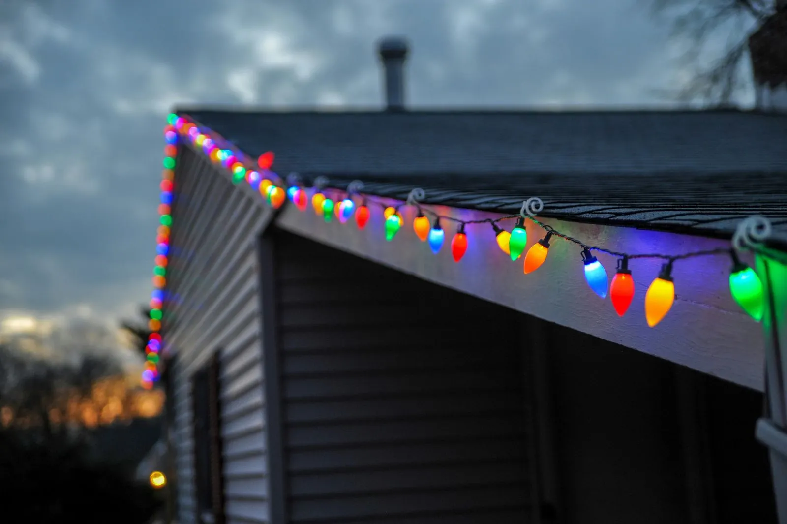 Multicolor C9 Christmas bulbs clipped along the roof edge of a house with vinyl siding at dusk