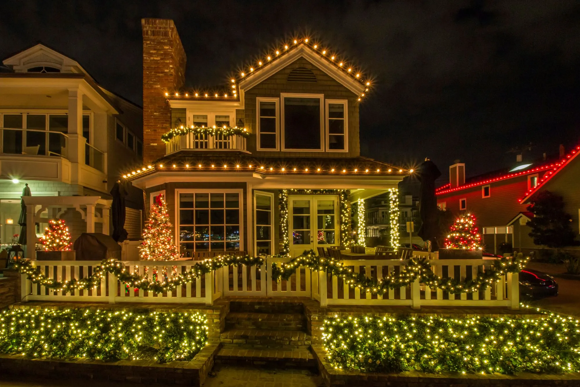 Shingle-sided two-story house outlined with warm white Christmas lights, lit garlands and a picket fence at night