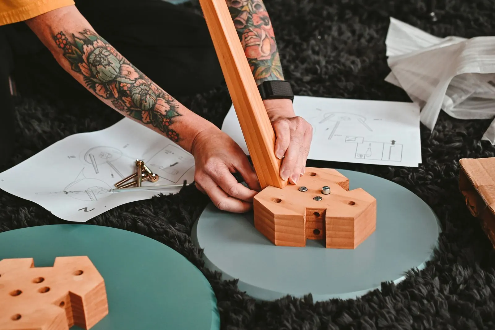Hands fitting a wooden leg into a table base on a rug, with the assembly instructions and screws beside it