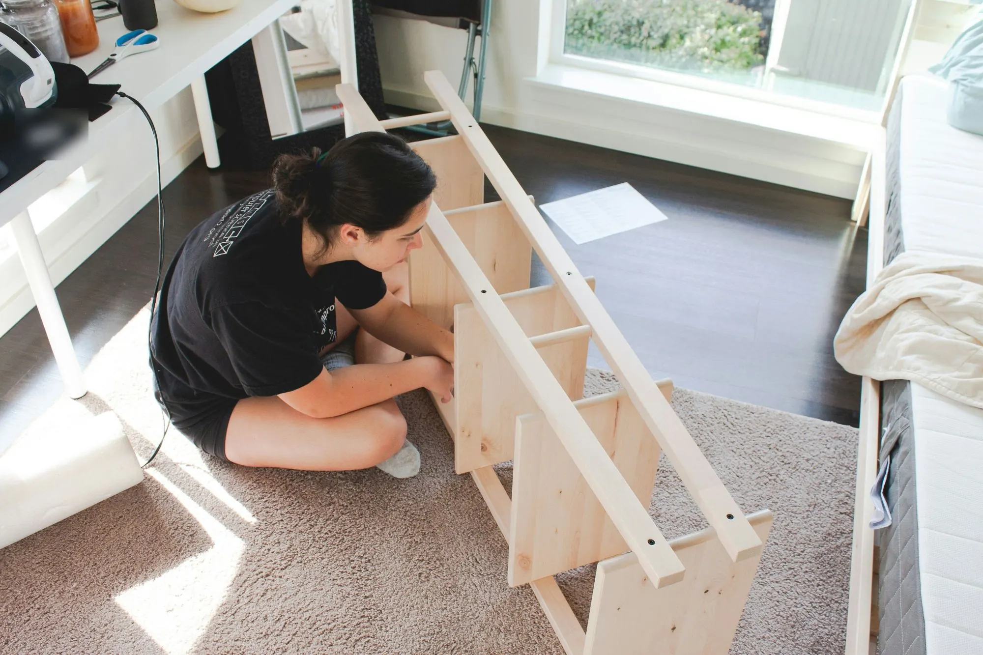 Woman sitting on a rug assembling an unfinished pine shelving unit in a sunny bedroom