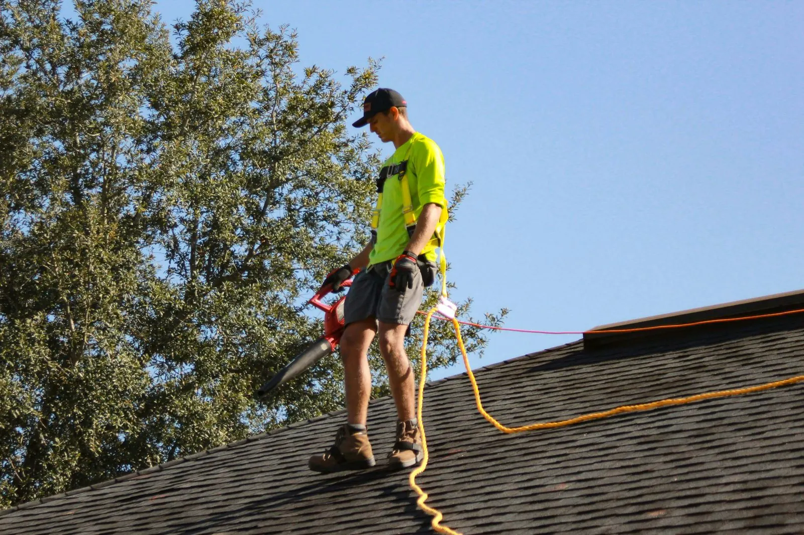 Worker in a safety harness and hi-vis shirt using a leaf blower to clear debris off a shingle roof